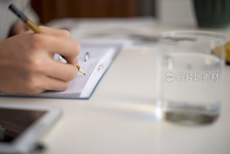 Unknown坐在一张白色的桌子旁，拿着一杯水和写字板，用铅笔在记事本上写字
