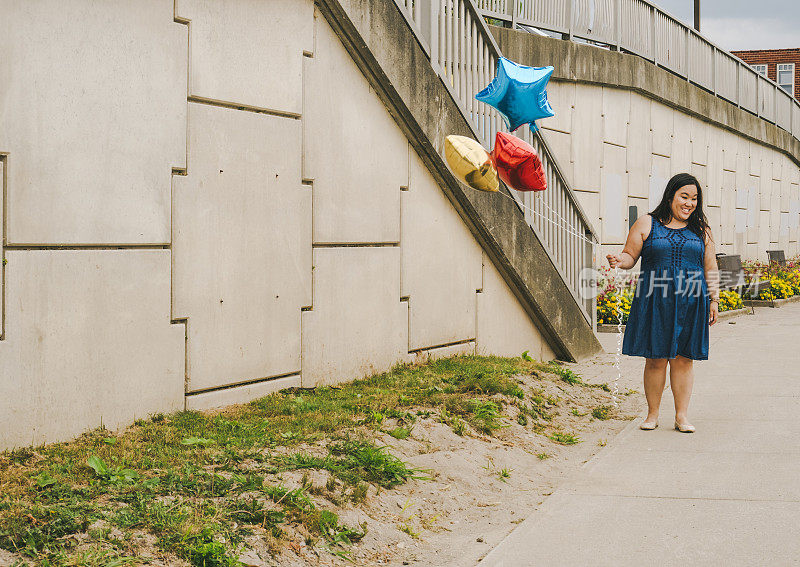 可爱的中国女人拿着三个气球沿着水泥墙走