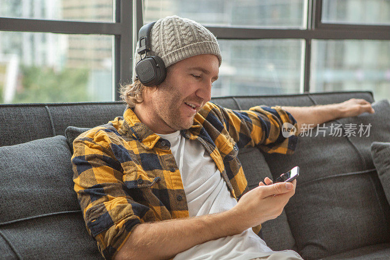 酷酷的年轻人坐在家里的沙发上使用智能手机和耳机