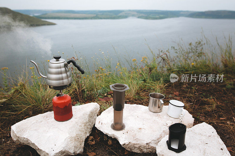 钢水壶沸腾的天然气primus，金属杯子和气压在悬崖上的背景湖，冲泡咖啡在露营。在户外野餐时制作热饮。在山上徒步旅行和远足