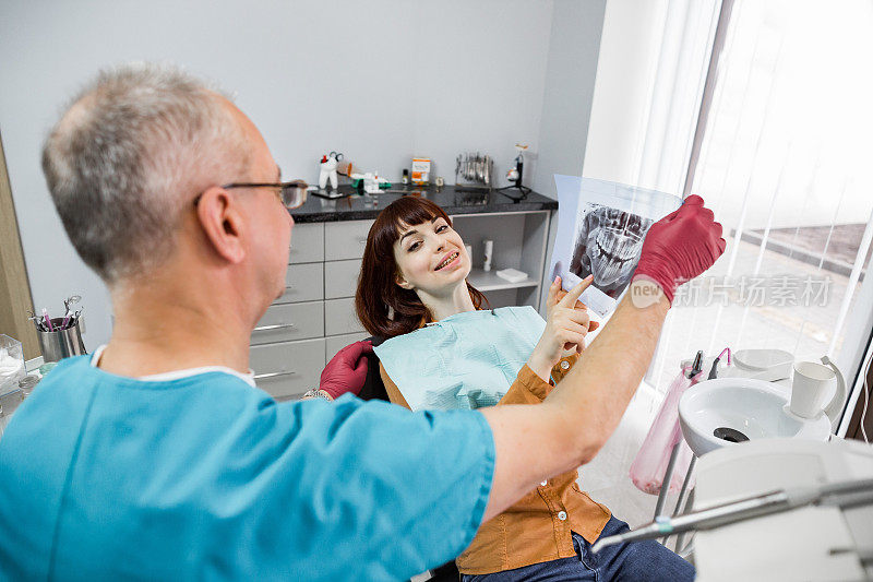 牙科诊所的资深男牙医拿着牙齿的x光片与女病人交谈，准备治疗。一位妇女在牙科诊所和她的牙医一起看x光片