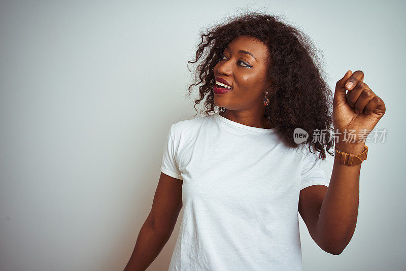 年轻的非洲裔美国妇女穿着t恤站在孤立的白色背景跳舞快乐和欢快，微笑着移动随意和自信听音乐