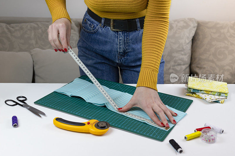 穿黄色衣服的女人测量织物。桌子上有缝被子的工具。拼布刀、剪刀、衬里剪垫、自锁、线、厘米胶带。缝纫用品