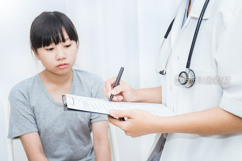 特写年轻的亚洲女孩在医生的诊所。亚洲女医生拿着笔检查检查检查清单纸。