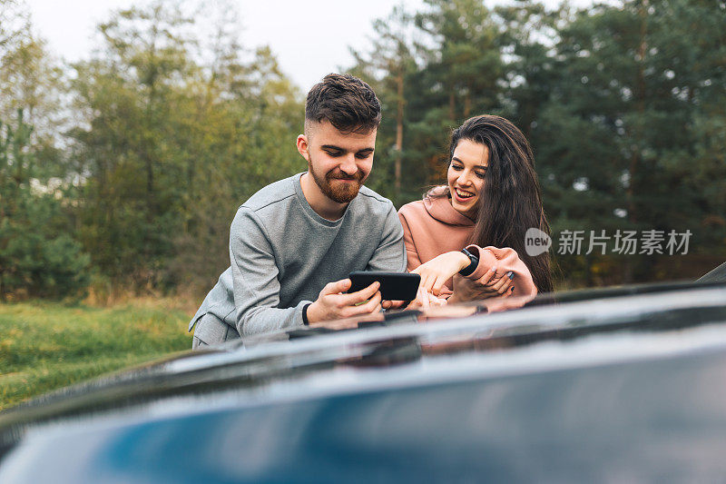 一对年轻夫妇倚靠在汽车引擎盖上，看着智能手机里一些令人愉快的东西