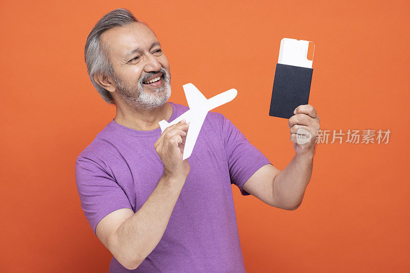 成熟的男人因为旅行而感到兴奋，拿着纸飞机，展示着机票和护照，孤立地站在橙色的背景下:-库存照片