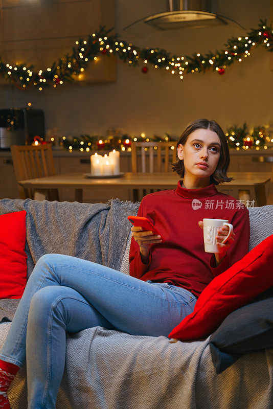为了过圣诞节，一位女士坐在装饰精美的灰色沙发上，若有所思地把目光移开。圣诞之夜，一位女士手里拿着一部智能手机和一个白色的杯子，杯子里放着热茶。