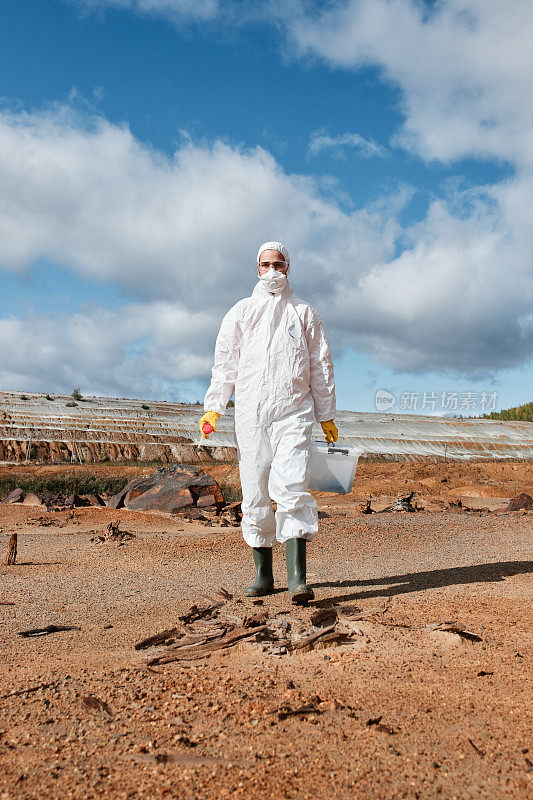 环境研究人员戴着手套、橡胶靴和防护服，带着装有土壤样本的容器在被污染的地面上行走