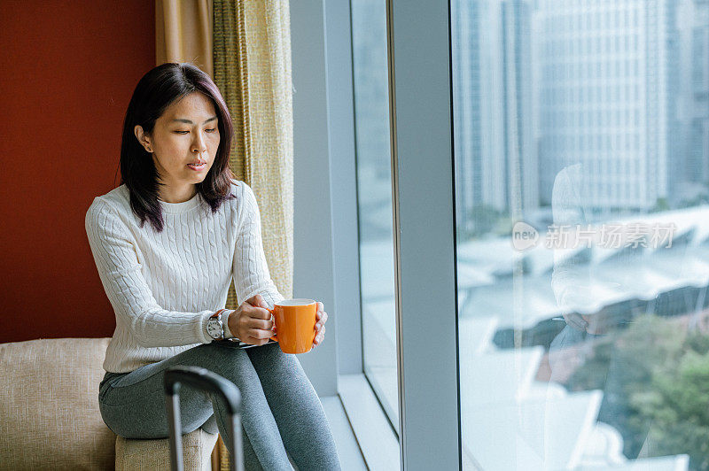 一名亚洲女游客在办理入住手续后，坐在窗边的沙发上，拿着咖啡在酒店房间里——独自旅行的概念