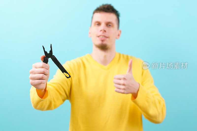 一名男子拿着一个多功能工具和钳子，在蓝色背景上竖起大拇指。口袋里。黑色的。刀片。设备。金属。多用途。实用程序。生存。开放。折叠。对象。处理