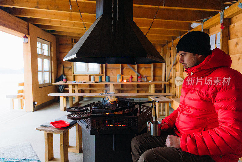 在挪威，一名身穿红色夹克的男子在真正的木制掩体里用火烹饪肉