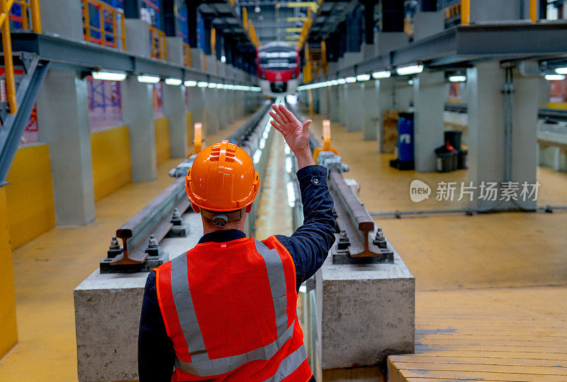 穿着安全制服的技术工人的背部向电气或地铁列车显示身体信号，并站在工厂工作场所的铁轨前。