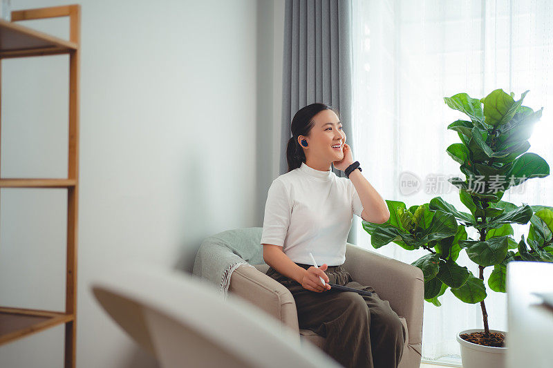 亚洲女性喜欢戴着耳机上网工作，在平板电脑上阅读数据，坐在家里的扶手椅上打电话。