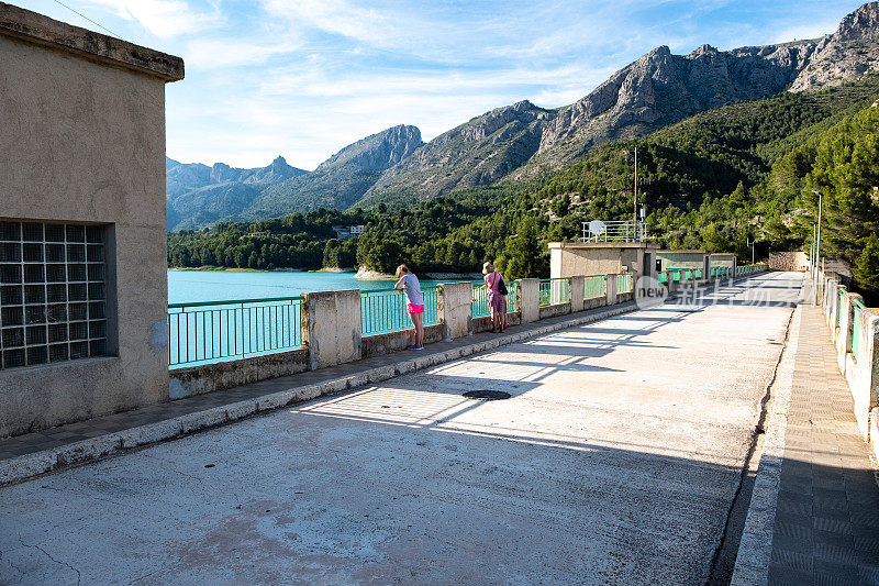 一群游客站在桥上欣赏美丽的风景，蓝色的湖泊和覆盖着森林的山脉。