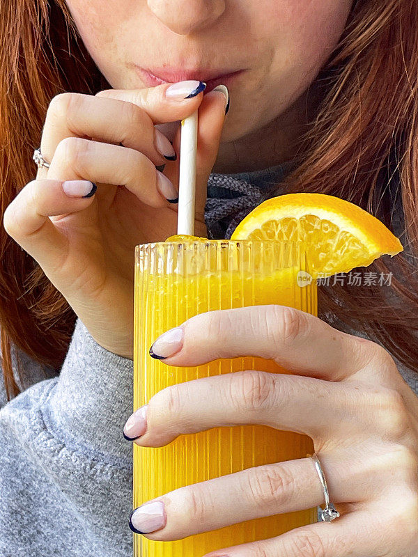 全画幅照片中，一个无法辨认的红发女人拿着一杯橙汁，里面有橙片和纸吸管，用吸管喝着橙汁，这是前景的焦点