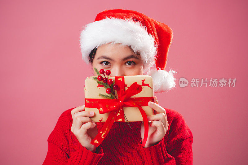 亚洲女人戴着圣诞老人的帽子，开心地微笑着，用礼盒遮住她的脸，孤立在粉红色的背景上。