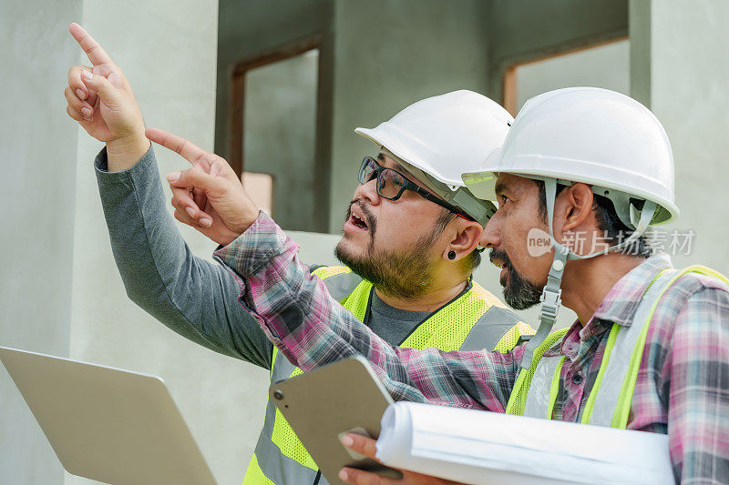 在建筑工地，一名男子穿着显眼的背心，手提电脑指向上方，与另一名戴着安全帽、手持平板电脑的男子交谈。建筑师和工程师监督房屋建设。