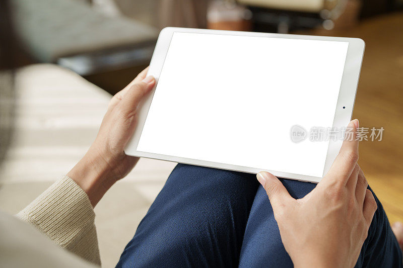 从肩膀的角度看，一位亚洲女性坐在沙发上，她的手正在使用电子平板电脑，同时在家工作，阳光温柔地照在她身上。