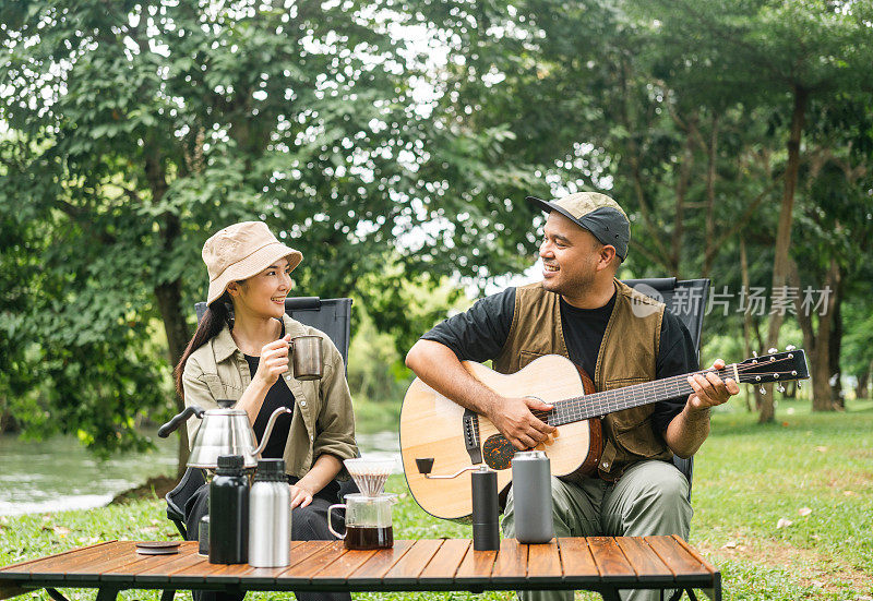 年轻的亚洲夫妇，男人和女人野餐坐在露营地户外花园。两个人在森林里露营，弹吉他，喝咖啡。旅游，放松，露营，度假，假日，周末的主题