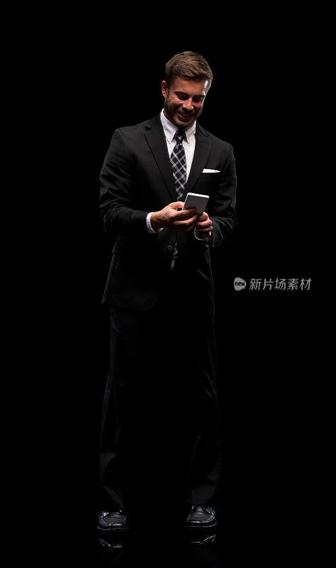 白人男性经理穿着商务装站在黑色背景前，使用智能手机