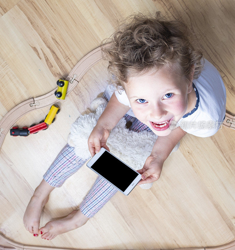 一个6岁的白人女孩坐在镶木地板上，手里拿着手机。前视图。可见的智能手机屏幕为复制空间