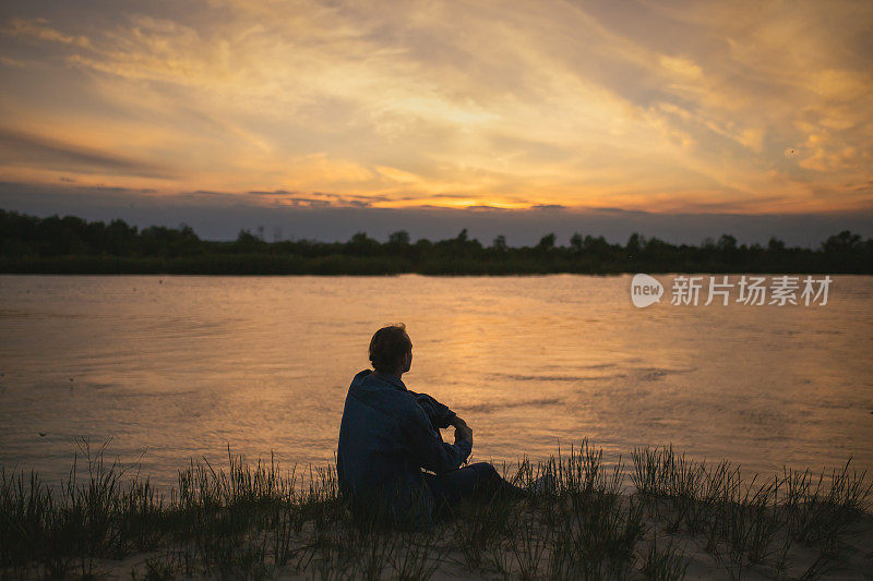 美丽的夏日夕阳背景上的男子剪影。橙色的天空。那家伙正坐在河边。人坐在河岸上