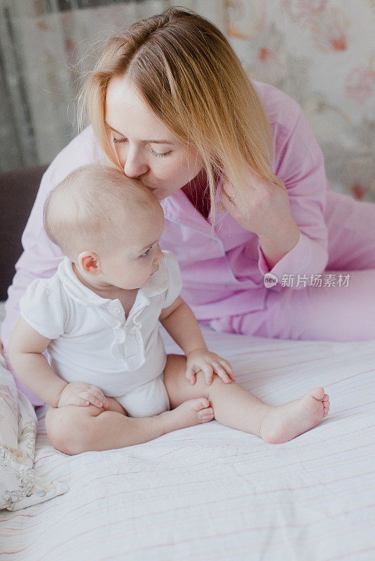 金发碧眼的母亲穿着粉红色的睡衣，亲吻着坐在她床上的婴儿