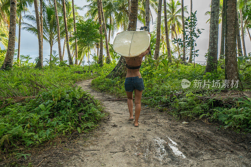 在菲律宾群岛的棕榈树下，手持长板的年轻女子冲浪女孩在海滩上走向大海。人们旅游度假运动概念。