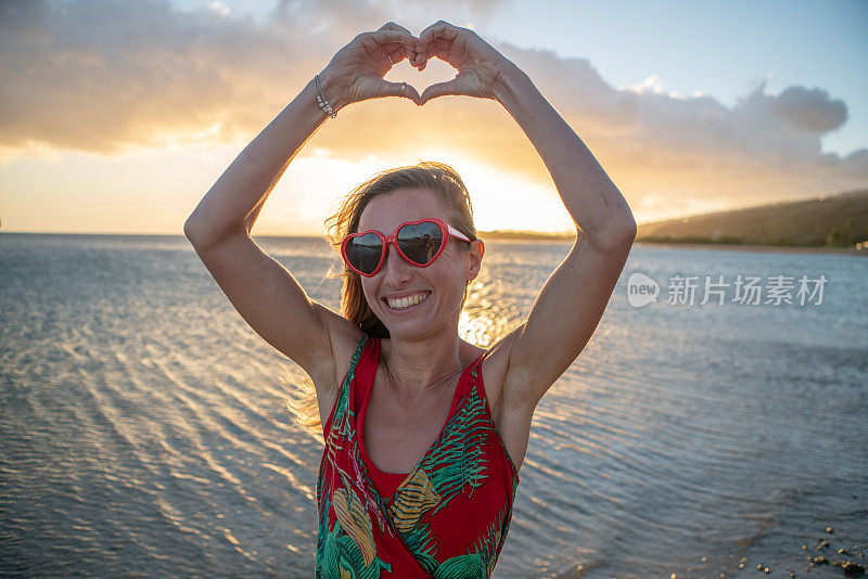 年轻女子的肖像戴心形红色太阳镜在美丽的海滩日落心形框架与手-圣瓦伦丁概念