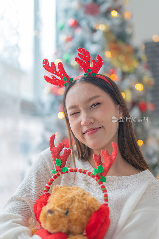 一个年轻美丽友好的亚洲女性女士的肖像照片可爱的圣诞驯鹿发带和柔软的玩具微笑愉快地在一个漂亮的装饰圣诞树的背景前