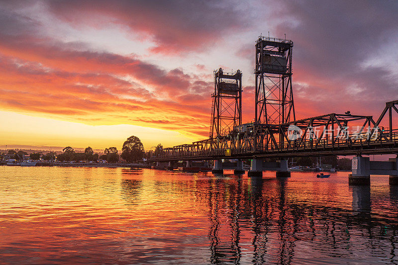 明亮戏剧性的金色天空在日落时反射在河水面上，有一座剪影的桥