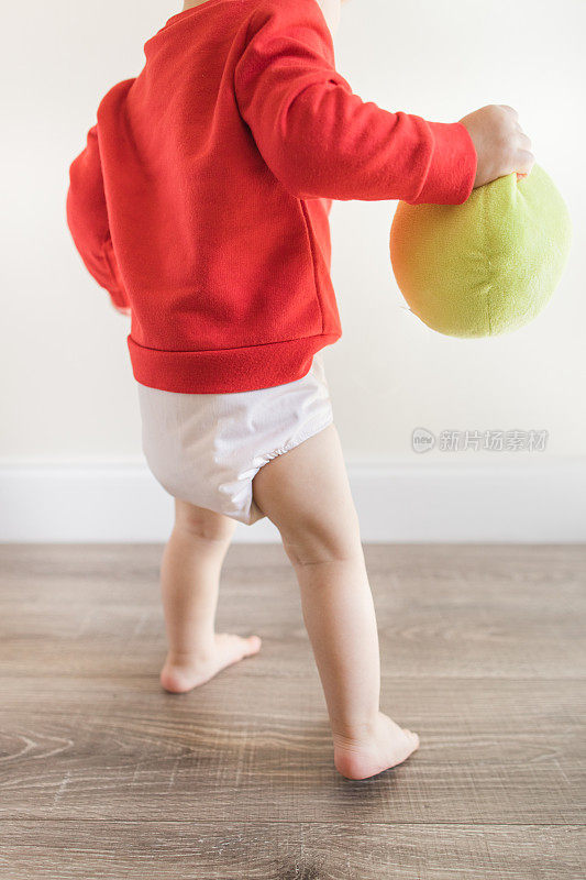 2023年2月的情人节，一个14个月大的可爱男婴赤脚站在木地板上，穿着写着“最小的情人节”的红色毛衣和裸色的布尿布，手里拿着一个绿色的球