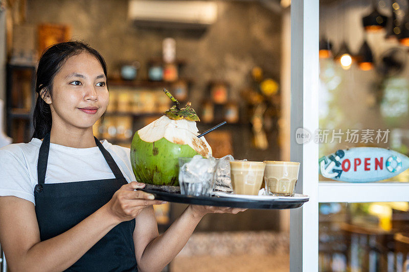 一个年轻美丽的女服务员在巴厘岛的一家餐厅工作，礼貌地为客人服务，端着美味的饮料，如卡布奇诺或椰奶在托盘上