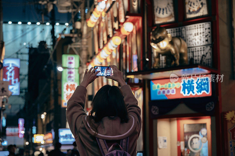 一位亚洲妇女正在日本大阪的堀林寺街观光。她正在用智能手机记录她的旅行记忆，并在社交媒体平台上分享。