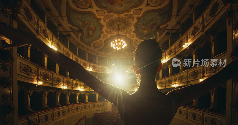 年轻的古典芭蕾舞女舞者站在聚光灯下的古典剧院舞台上谢幕的电影后视图。优雅的表演者接受观众对她的才华的掌声
