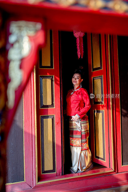 身穿泰国兰纳传统服饰的年轻亚洲女子站在红色的门边，古宅的古风也面带微笑地望向左边。