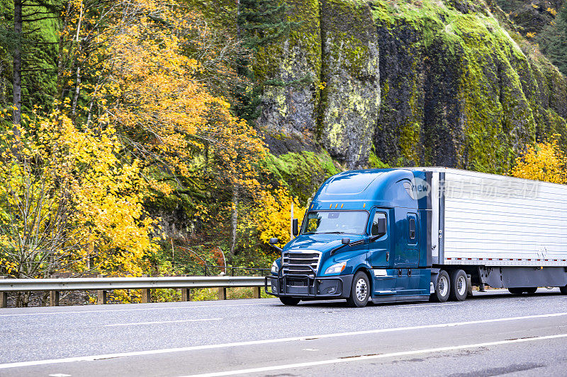深蓝大钻机专业半挂车运输货物在冰箱半挂车行驶在秋天的道路上与山石和黄色的树在一边