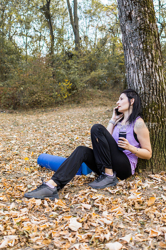 秋天，一位中年妇女在树叶环绕的森林里练完瑜伽后正在使用手机。她有锻炼的器材。