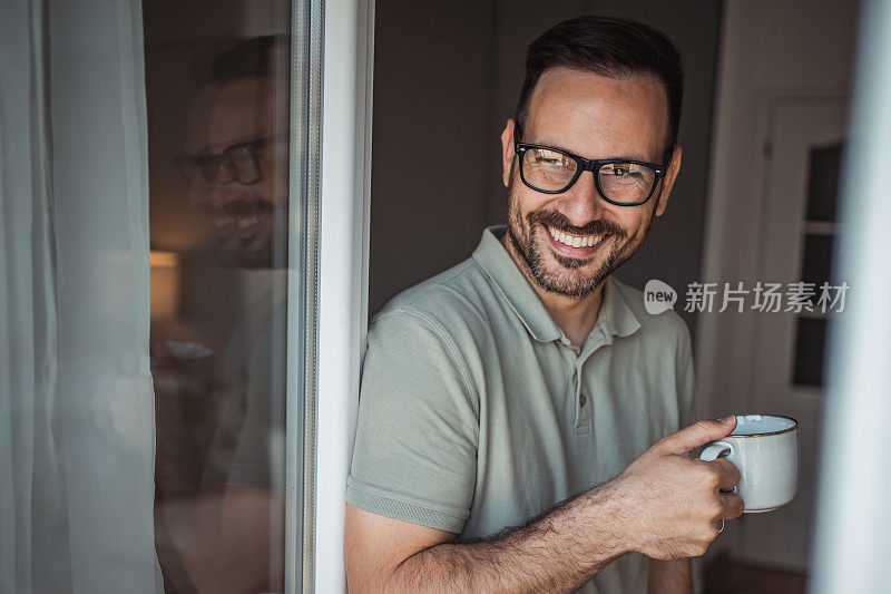 一位成年男子端着一杯咖啡，面带微笑地站在公寓的阳台门口