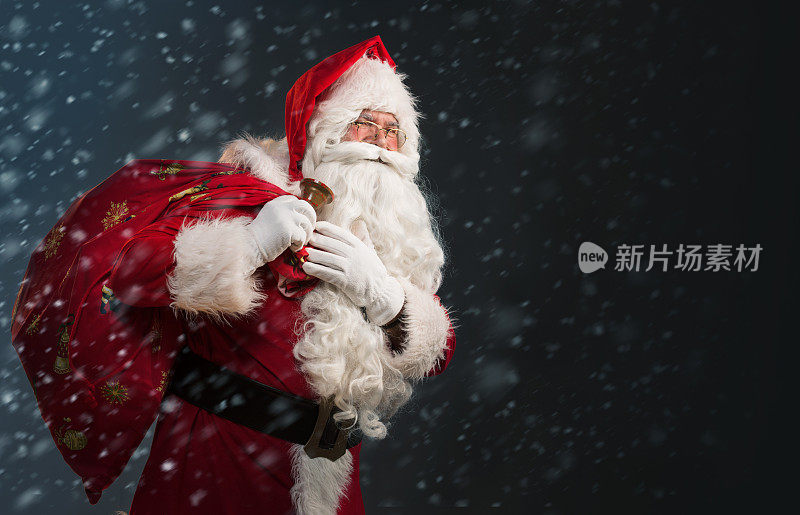 圣诞老人拿着一个装着礼物的袋子，在一个有雪的黑暗背景上摇铃