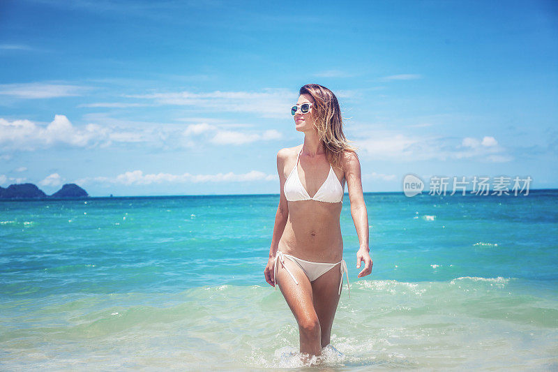 美丽的年轻女子穿着白色比基尼走在热带海滩上