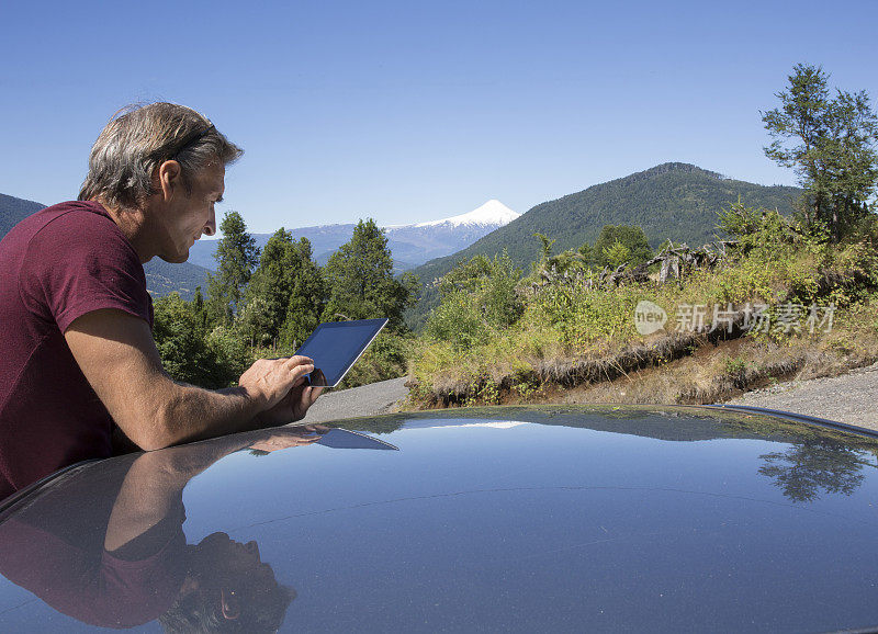 成熟的男人在车顶上使用数码平板电脑，后面是火山
