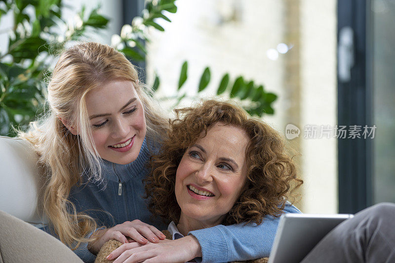 年长的妈妈和成年的女儿在沙发上用数码平板电脑。你看我，我看你。