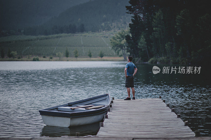 在一个美丽的湖上，一个人的手在臀部站在一个木制的码头上，与一个系泊的划艇在大自然中