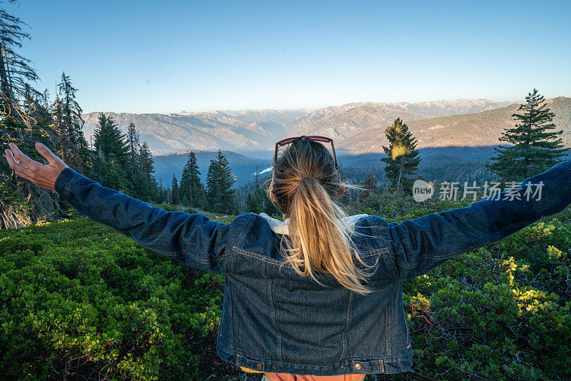 年轻女子站在森林里，张开双臂看着美丽的山谷日落