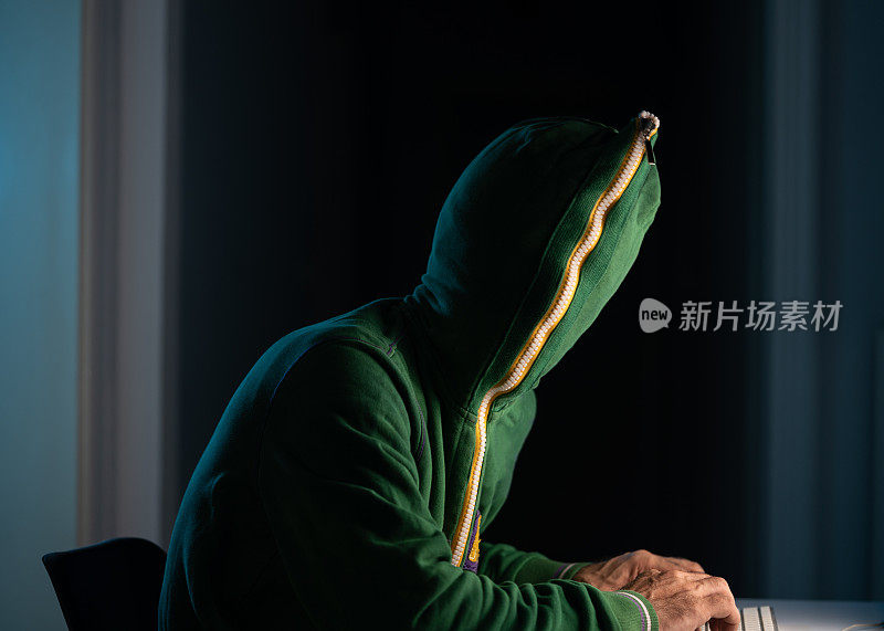 一名穿着带帽拉链衬衫和面具的男子在黑暗中使用笔记本电脑入侵