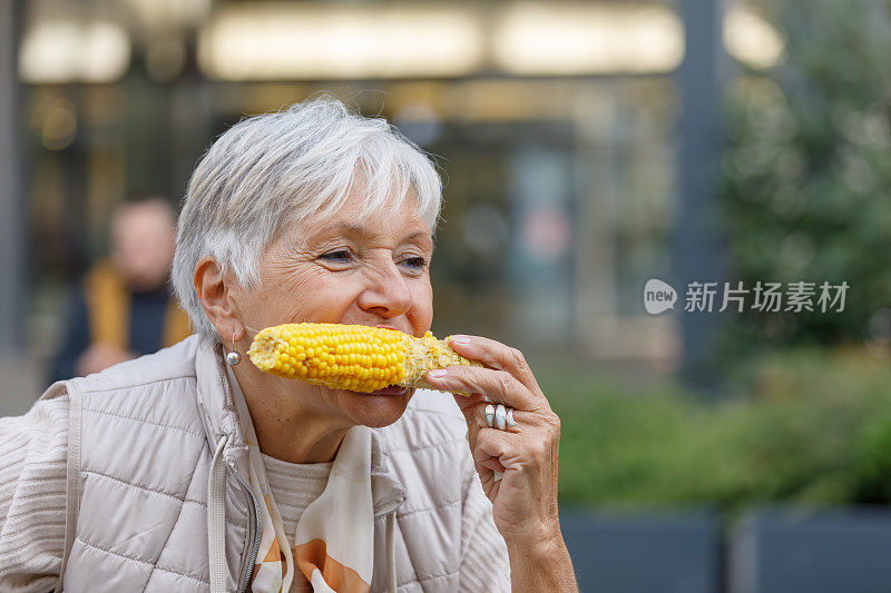一位老妇人一边吃着煮熟的玉米棒，一边在城里散步。