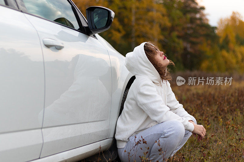 一个穿着白色连帽衫和运动裤的漂亮年轻女子坐在一辆白色汽车旁边，在一个温暖的秋日里，映衬着黄色树叶的背景。有选择性的重点