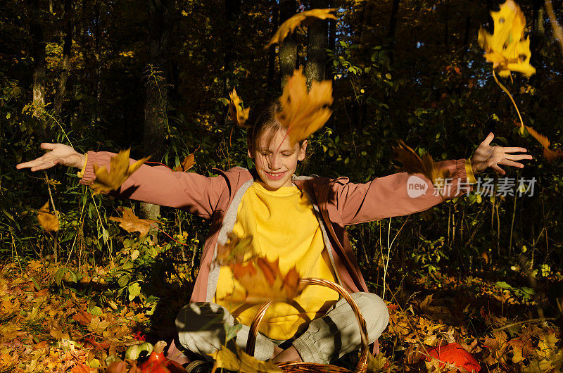 快乐的小女孩穿着黄色的运动衫和满是灰尘的淡紫色外套，坐在秋天的森林里扔着黄叶。