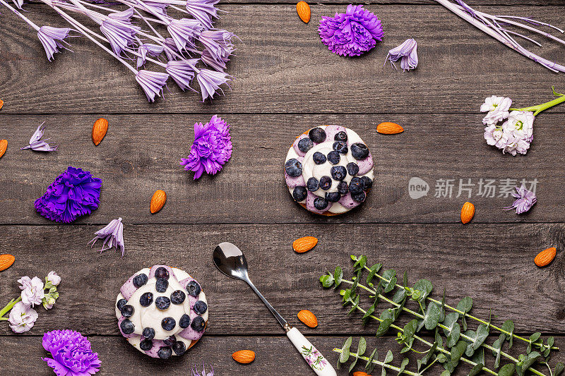 蓝莓格拉汉姆芝士蛋糕上点缀着新鲜的浆果，勺子放在深色的木质背景上，点缀着杏仁、紫色的花朵和小树枝。平的。